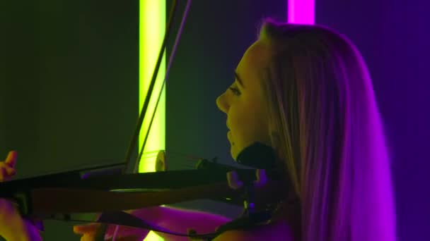 Veduta laterale di un'adorabile violinista che suona un violino originale in legno. La bionda fa un recital dal vivo a una festa tra luci al neon luminose. Chiudete. Rallentatore. — Video Stock