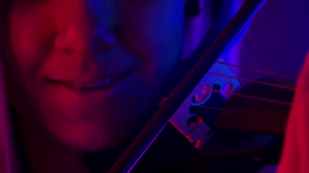 Részletes felvétel egy fa hegedűről és masniról. Egy profi hegedűművész klasszikus szimfóniát játszik fényes neonfények hátterében. Női arc és szerszám közelről. Lassú mozgás.. — Stock videók