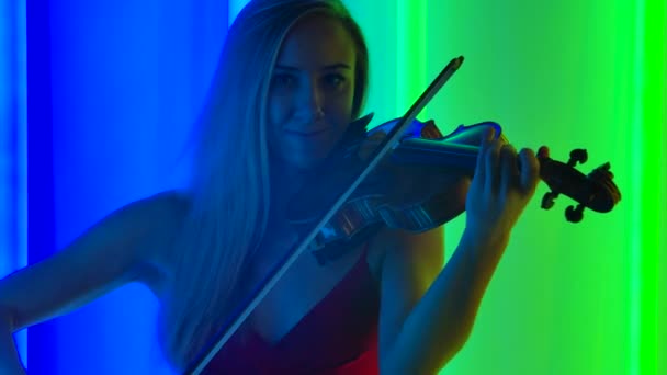 En ung kvinnlig violinist framför en melodi av klassisk instrumentalmusik på fiolen. Söt blondin med långt hår i en studio med ljusa neonljus. Närbild. Långsamma rörelser. — Stockvideo