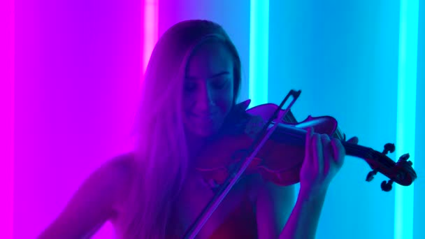 En ung kvinnlig violinist framför en melodi av klassisk instrumentalmusik på fiolen. Söt blondin med långt hår i en studio med ljusa neonljus. Närbild. Långsamma rörelser. — Stockvideo