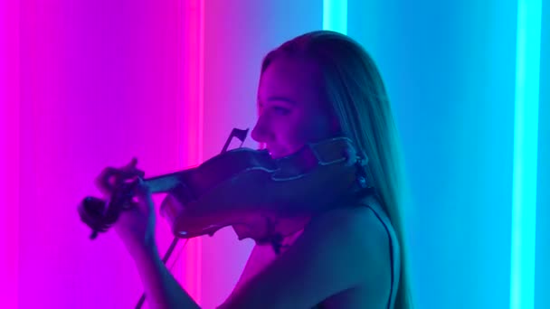 Zijaanzicht van een vrouwelijke muzikant met lang haar die vioolsymfonie speelt. Een jonge artiest treedt op op een feestelijk feest tegen de achtergrond van felle neonlichten. Sluit maar af. Langzame beweging. — Stockvideo