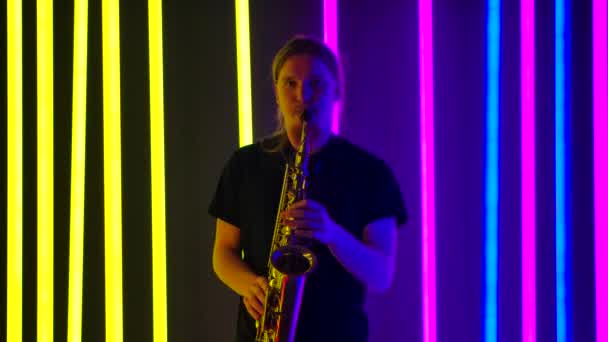 En ung man i svart T-shirt spelar saxofon i en mörk studio mot bakgrund av starka neonljus. Långsamma rörelser. — Stockvideo