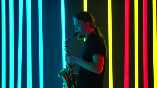 Sidovy av en ung snygg man med långt hår spelar saxofon mästerligt. Musikern framför en livekonsert i en mörk studio mot bakgrund av starkt neonljus. Långsamma rörelser. — Stockvideo