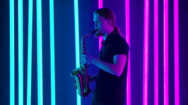 Sidovy av en ung snygg man med långt hår spelar saxofon mästerligt. Musikern framför en livekonsert i en mörk studio mot bakgrund av starkt neonljus. Långsamma rörelser. — Stockvideo