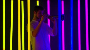 Genç yakışıklı adam trompet çalmaktan hoşlanıyor. Stüdyodaki parlak neon ışıklar arasında müzikal performans. Gece hayatı ve eğlence konsepti. Yavaş çekim.