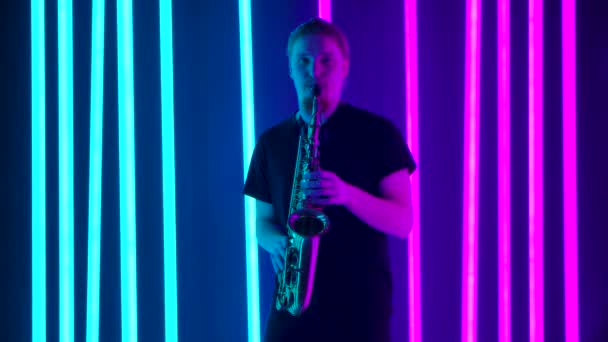 En ensam manlig saxofonist uppträder i en mörk ateljé mot bakgrund av starkt neonljus. En man i svart T-shirt spelar blåsinstrumentet professionellt. Långsamma rörelser. — Stockvideo