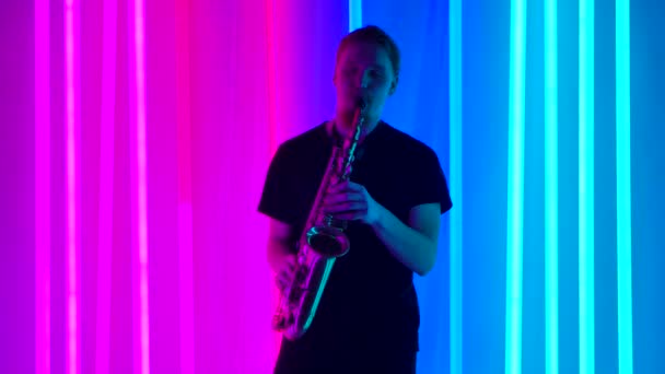 Un saxofonista macho solista actúa en un estudio oscuro contra el telón de fondo de brillantes luces de neón. Un hombre con una camiseta negra toca el instrumento de viento profesionalmente. Movimiento lento. — Vídeos de Stock