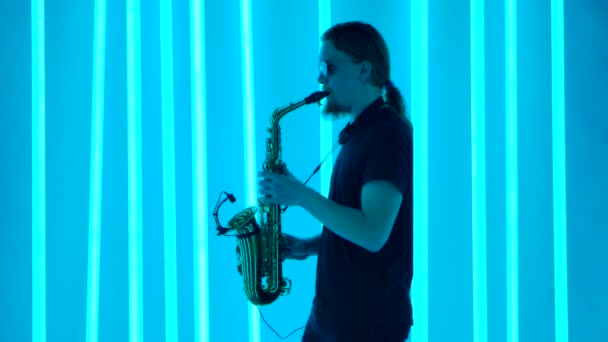 Boční pohled na saxofonistu se stylovými slunečními brýlemi, který si užívá hraní na hudební nástroj. Člověk hraje klasickou hudbu v tmavém studiu na pozadí jasně modrých světel. Zpomalený pohyb. — Stock video