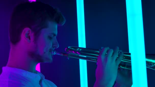 Sidovy av en manlig musiker som spelar trumpet entusiastiskt. Musikalisk prestanda bland starkt neonljus. Mässing blåsinstrument i händerna på en man närbild. Långsamma rörelser. — Stockvideo
