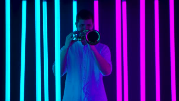 Live optreden van een muzikant die trompet speelt. Een man in een wit shirt speelt een jazzmelodie in een donkere studio tegen de achtergrond van fel neonlicht. Langzame beweging. — Stockvideo