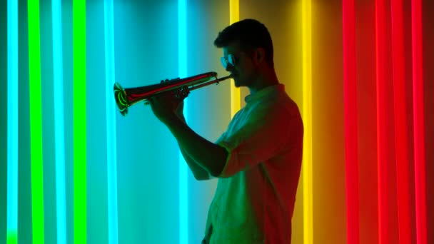 サングラスの若いスタイリッシュな男が熱心にトランペットを演奏します。明るい色のネオンでレトロなスタイルの音楽パーティー。スローモーション. — ストック動画
