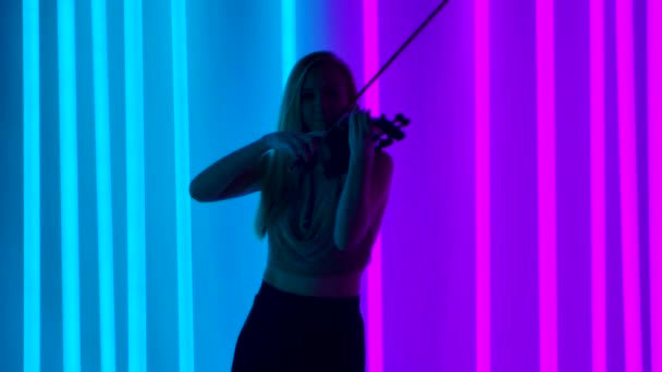 Silhueta de jovem loira atraente toca violino entre as luzes de néon brilhantes. Uma mulher toca as cordas dos arcos enquanto toca música clássica. Movimento lento. — Vídeo de Stock