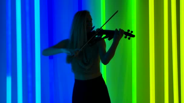Genç ve çekici sarışının silueti parlak neon ışıklar arasında keman çalıyor. Bir kadın klasik müzik çalarken yayların tellerine dokunur. Yavaş çekim. — Stok video