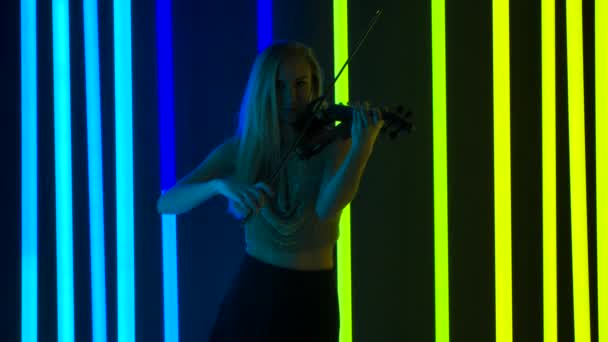 Glimlachende vrouwelijke muzikant speelt meesterlijk viool in de studio. Een blondine in een glanzend topje pronkt met haar dapperheid tegen een heldere neon achtergrond. Langzame beweging. — Stockvideo