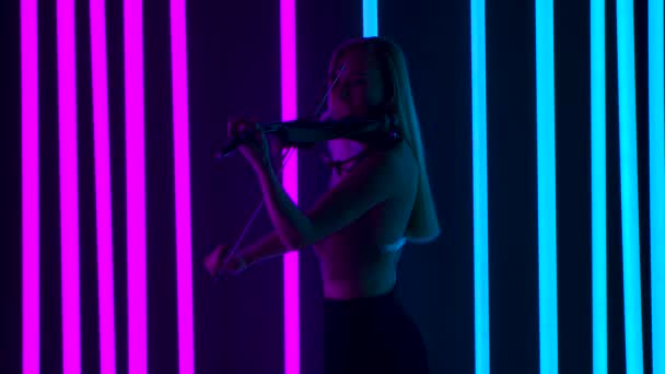 Vista lateral de una adorable violinista tocando un violín de madera. La rubia realiza un recital en vivo en una fiesta en medio de brillantes luces de neón. Movimiento lento. — Vídeo de stock