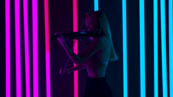 Vista lateral de una adorable violinista tocando un violín de madera. La rubia realiza un recital en vivo en una fiesta en medio de brillantes luces de neón. Movimiento lento. — Vídeo de stock