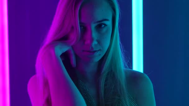 Egy fiatal, szexi nő portréja, aki a kamera előtt pózol. Szőke, hosszú hajjal, sötét stúdió háttérrel, világos, sokszínű neonfényekkel. Lassú mozgás. Közelről.. — Stock videók