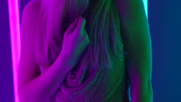 Retrato de uma jovem mulher sexy posando na frente da câmera. Loira com cabelos longos em um fundo de estúdio escuro com luzes de néon multicoloridas brilhantes. Movimento lento. Fechar. — Vídeo de Stock