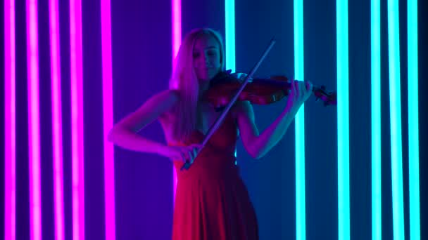 En ung kvinnlig violinist framför en melodi av klassisk instrumentalmusik på fiolen. En söt blondin i en röd klänning uppträder på en musikalisk kväll mitt i ljusa neonljus. Långsamma rörelser. — Stockvideo