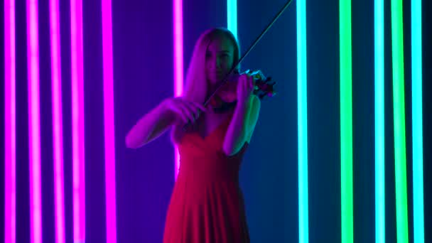 Młoda skrzypaczka gra na skrzypcach melodię klasycznej muzyki instrumentalnej. Ładna blondynka w czerwonej sukience występuje w muzycznym wieczorze pośród jasnych neonów. Zwolniony ruch. — Wideo stockowe