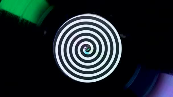 Ovanifrån av en svart vinyl skiva med en hypnotisk etikett snurrar mot en bakgrund av blinkande färgade neonljus. Retro skivspelare närbild i slow motion. — Stockvideo