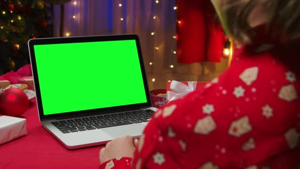 Bekijk van achteren een vrouw op een laptop met een groen scherm chroma sleutel op de achtergrond van een interieur ingericht voor oudejaarsavond. Online communicatie via videochat. Sluit maar af. Langzame beweging. — Stockvideo