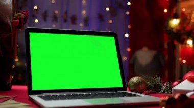 Yeşil ekran krom anahtarı olan dizüstü bilgisayar Noel Baba 'nın yanındaki kırmızı masada duruyor. Hediyeler ve oyuncaklar. Noel dekorlu, arka planda bulanık ışıklar olan ev odası. Model monitörü. Kapatın. Yavaş çekim.
