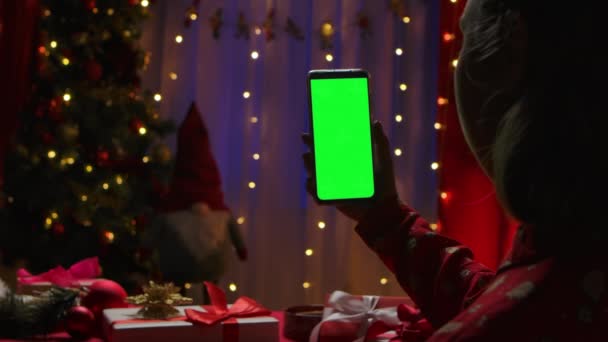 Vrouw houdt in haar hand een smartphone met groene scherm chroma sleutel op de achtergrond van een interieur ingericht op oudejaarsavond. Online communicatie via videochat. Sluit maar af. Langzame beweging. — Stockvideo