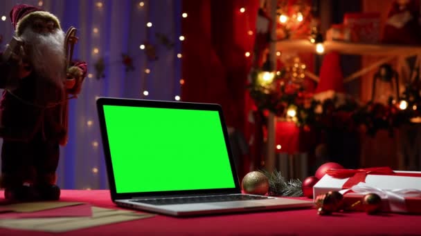 Laptop con chiave cromatica schermo verde si trova sul tavolo rosso accanto a Babbo Natale, regali e giocattoli. Camera di casa con decorazioni natalizie e luci sullo sfondo sfocato. Monitor Mockup. Chiudete. Rallentatore. — Video Stock