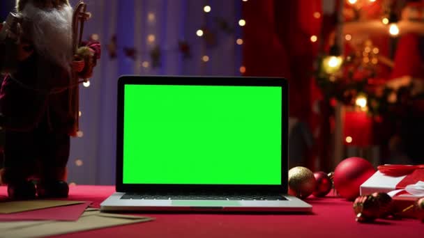 Laptop con chiave cromatica schermo verde si trova sul tavolo rosso accanto a Babbo Natale, regali e giocattoli. Camera di casa con decorazioni natalizie e luci sullo sfondo sfocato. Monitor Mockup. Chiudete. Rallentatore. — Video Stock