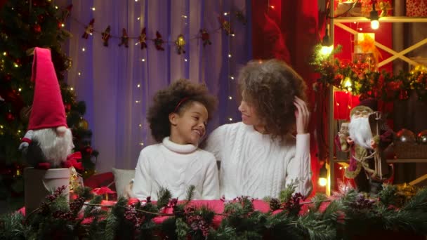 アフリカ系アメリカ人の母親と彼女の小さな娘は歌を歌い、楽しんでいます。クリスマスツリーの近くに座っている女性と女の子は、お祝いの夜のために飾られた家の部屋の背景を背景に. — ストック動画