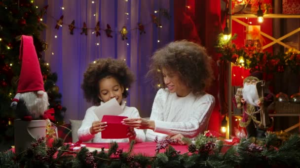 幸せなアフリカ系アメリカ人の母親と小さな娘はサンタクロースに手紙を書き、喜んで封筒に詰めます。幸せな家族の肖像画、新年のお祝いのコンセプト. — ストック動画