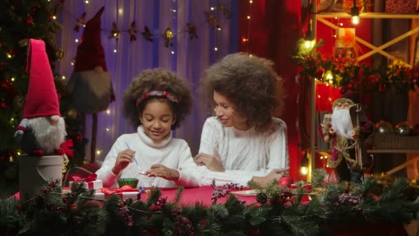 Ein kleines afroamerikanisches Mädchen bemalt ein Weihnachtsspielzeug. Mutter und Tochter sitzen vor der Kulisse des heimischen Zimmers, dekoriert für die festliche Nacht. Glückliches Familienporträt, Konzept für die Neujahrsfeier. — Stockvideo