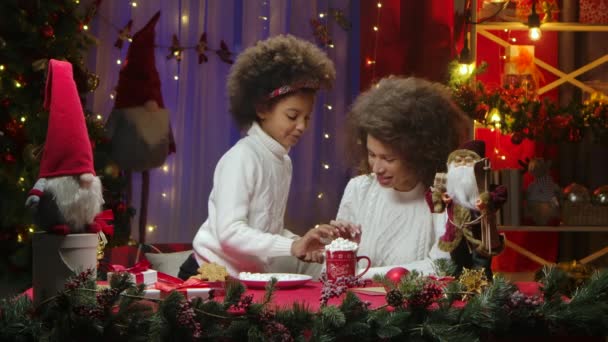 Mamma afroamericana e figlioletta guarniscono una tazza di bevanda calda con panna montata e marshmallow. Felice ritratto di famiglia, concetto di celebrazione di Capodanno. — Video Stock