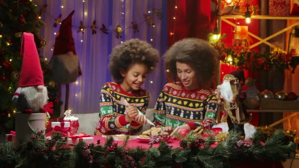 Niña afroamericana deliciosamente decora galletas caseras de Navidad con crema. Feliz retrato familiar, concepto de celebración de Año Nuevo. — Vídeo de stock