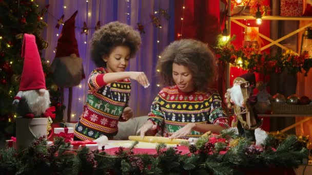 Maman afro-américaine et sa fille en chandails de vacances lumineux s'amuser et déployer la pâte pour les cookies maison de vacances. Portrait de famille heureux, concept de célébration du Nouvel An. — Video