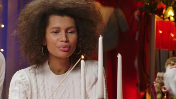 Okouzlující Afroameričanka v bílém svetru svítí bílé sváteční svíčky. Žena sedí na pozadí domácí místnosti, zdobené pro vánoční svátky. Zavřít. — Stock video