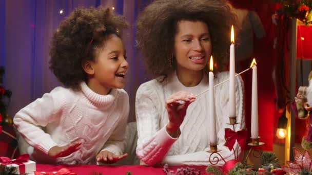 Афро-американская мама и маленькая дочь в белых свитерах зажигают свечи от восторженных эмоций. Счастливый семейный портрет, концепция празднования Нового года. Закрыть. — стоковое видео