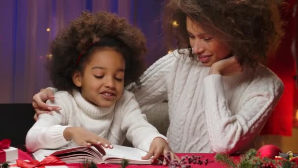 Beyaz kazaklı Afrikalı anne ve küçük kızı bir kitap okuyup mutlu mesut sarılıyorlar. Mutlu aile fotoğrafları, yeni yıl kutlaması konsepti. Kapat.. — Stok video