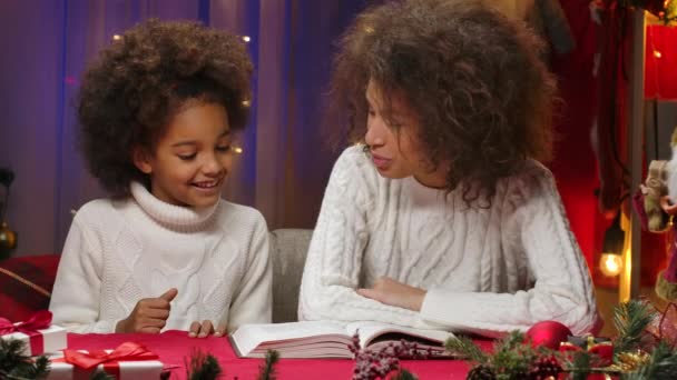 Afrikansk amerikansk mor og lille datter i hvide trøjer læser en bog og kæler lykkeligt. Happy familie portræt, nytår fest koncept. Tæt på. – Stock-video