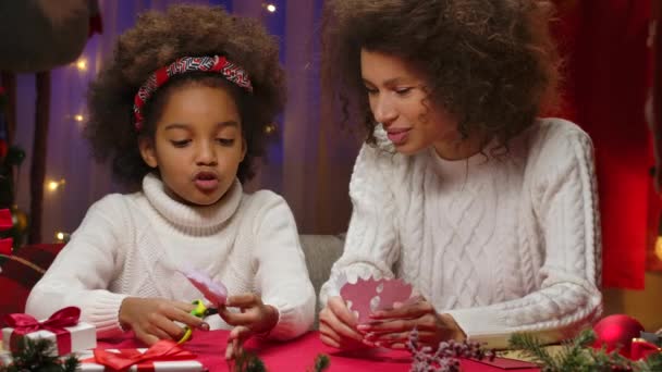 Little African American girl cuts snowflakes from paper. Mom and daughter in white sweaters sitting against the background of the home room, decorated for the Christmas holidays. Close up. — Stock Video
