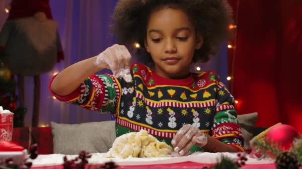 Little African American girl in a New Years print sweater prepares dough for homemade cookies. Child on the background of the kitchen decorated for the Christmas holidays. Slow motion. Close up. — Stock Video