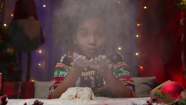 Petite fille afro-américaine dans le pull imprimé Nouvel An s'amuse, souffle de la farine de ses mains et prépare de la pâte pour les biscuits faits maison. Joyeux Noël concept. Au ralenti. Gros plan. — Video