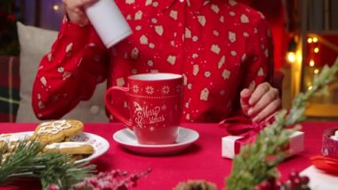 Kırmızı pijamalı ve yeni yıl baskısı olan bir kadın bir fincan sıcak içeceği kremayla süslüyor. Noel süslemeleriyle süslenmiş bir odanın arka planındaki kadın ellerini kapat. Yavaş çekim.