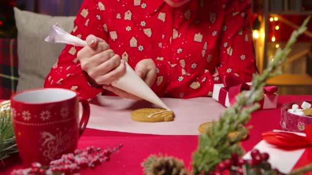 Τα γυναικεία χέρια στολίζουν τα παραδοσιακά χριστουγεννιάτικα μπισκότα με κερασάκι. Γυναίκα με κόκκινες πιτζάμες με εορταστική εκτύπωση στο φόντο της κουζίνας διακοσμημένη για τις διακοπές της Πρωτοχρονιάς. Κλείσε. Αργή κίνηση. — Αρχείο Βίντεο