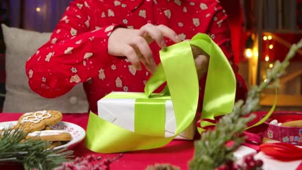 Mujer en pijama rojo con estampado de Año Nuevo envuelve una caja de regalo y ata un lazo. Primer plano de las manos femeninas en el fondo de una habitación decorada con decoraciones y regalos de Navidad. Movimiento lento. — Vídeos de Stock