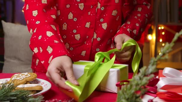 Жінка в червоній піжамі з новорічним принтом обмотує подарункову коробку і зав'язує лук. Крупним планом жіночі руки на тлі кімнати, прикрашеної різдвяними прикрасами та подарунками. Повільний рух . — стокове відео