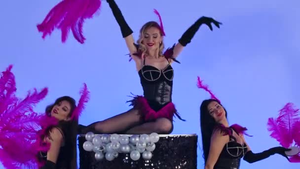 Jeunes femmes sexy en body en cuir avec des fans de plumes violettes dansant sur un gâteau noir. Trois danseurs burlesques posant sur fond bleu clair en studio. Ferme là. Mouvement lent. — Video