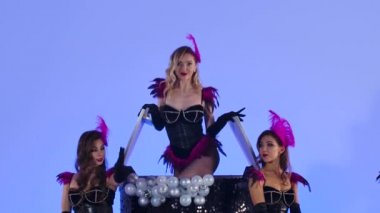 Şık, seksi striptizci büyük, siyah bir pastanın içinden çıkıyor. Stüdyoda açık mavi arka planda siyah elbiseli güzel kadınlar poz veriyor. Kapatın. Yavaş çekim.