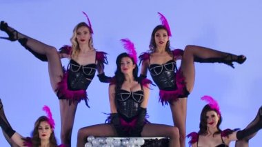 Mor tüylü siyah elbiseli seksi genç kadınlar pastanın üzerinde dururken dans ediyorlar. İzole edilmiş mavi arka planda poz veren bir grup kabare dansçısı. Ağır çekim. Kapat..